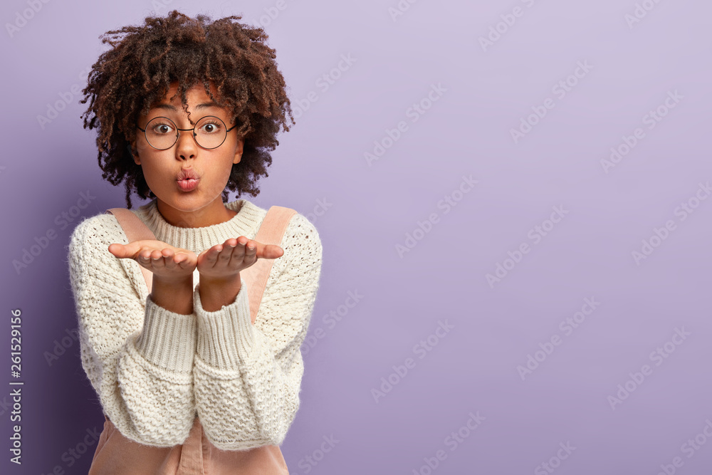 Indoor shot of lovely beautiful mixed race female blows air kiss, stretches hands near mouth, keeps lips folded, dressed casually, stands over purple background with free space for your promotion Indoor shot of lovely beautiful mixed race female blows air kiss, stretches hands near mouth, keeps lips folded, dressed casually, stands over purple background with free space for your promotion