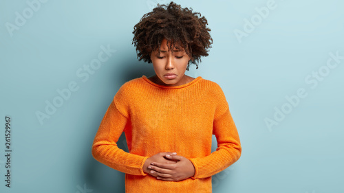 Foto Discontent Afro American female suffers from abdominal stomach pain, touches belly with both hands, looks down, wears orange jumper, has menstruation, curly haircut, models over blue background