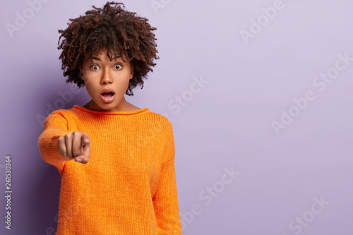 Photo of surprised startled emotional curly woman points fore finger straight at camera  opens mouth from shock  demonstrates something in front  wears casual clothes  models over purple background