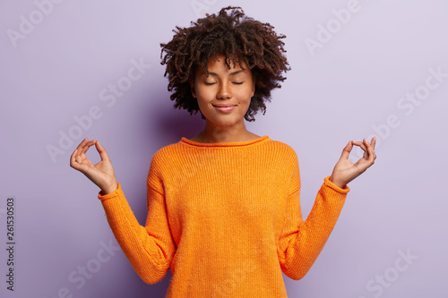 Pleasant looking calm woman meditates indoor, holds hands in mudra gesture, has charming smile, closed eyes, wears orange clothes, models over purple background. Hand gesture. Meditation concept
