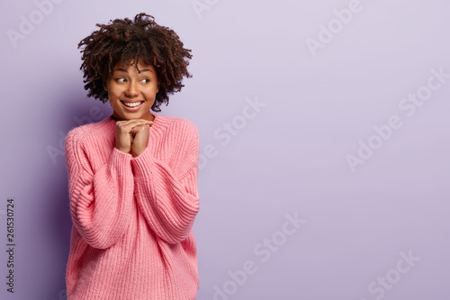 Waist up shot of good looking smiling teenage girl keeps both hands together under chin, focused away, feels shy, gets compliment from stranger, looks with delighted expression. Human emotions