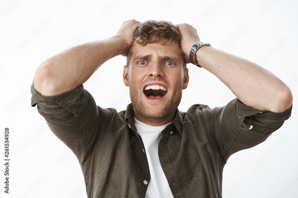 Intense shocked guy in stupor with blue eyes and fair hair holding hands on head yelling, screaming in panic, freaking out, standing stunned and insecure over gray background scared and frightened