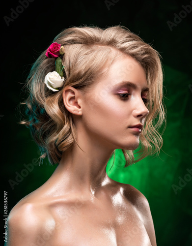 Beautiful young blonde girl with naked shoulders  flowers braided into her hair and oily body poses on a dark green gradient background. Clean  healthy skin. Nude make-up. Close up