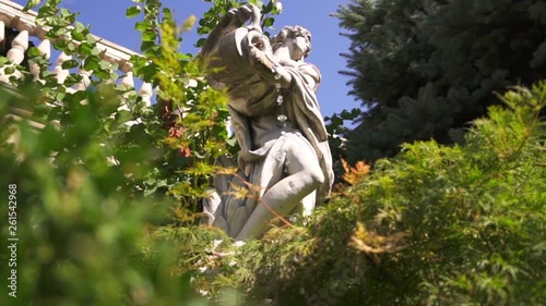 Classical gypsum figure holds amphora with water that drops on grass in front of baluster terrace in a green garden on clear blue sky background. Sculpture garden with trees, fir-trees and bushes.