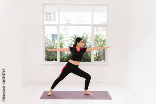 Young attractive woman practicing yoga, standing in Warrior two exercise, wearing sportswear, indoor full length near window