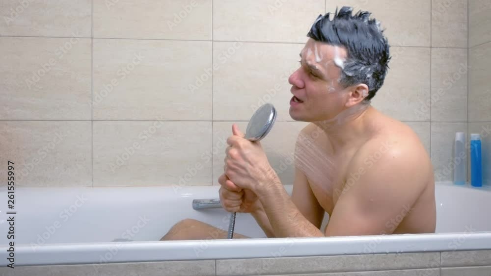 Happy man with Mohawk shampoo hairs is singing while he is washing in bathtub. Side view.