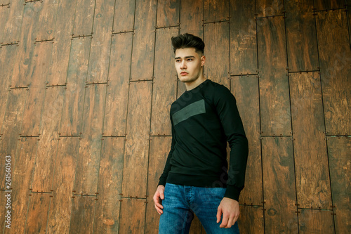 young brunette guy and model with a fashionable haircut and styling in a black jacket on a wooden background with a place for the inscription © Карина Желнина