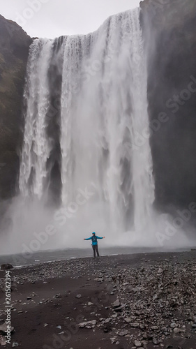 A young man wearing blue jacket standing under the massive waterfall with his arms wide spread. Moody atmosphere. Pure happiness and enjoyment. Gesture of freedom. Beauty of the nature