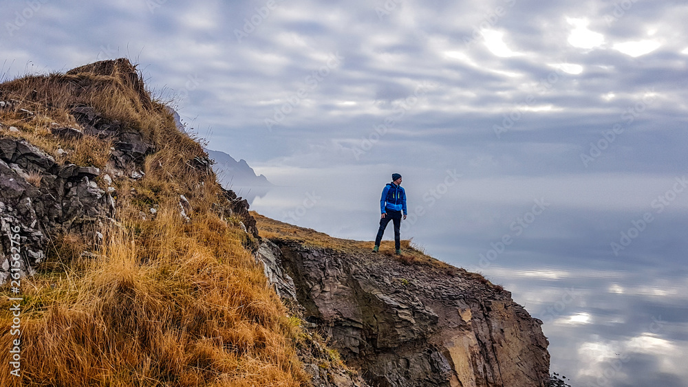 A young man wearing blue jacket stands at the edge of a cliff, looking into the sea. Tall mountains hiding in the mist behind him. Sharp cliff edges surround the man. Skyline blending with the water.