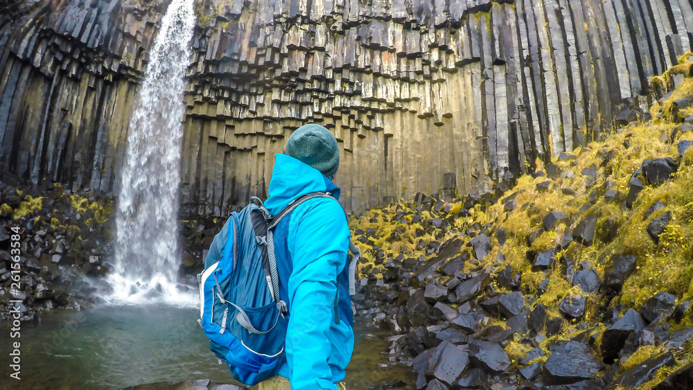 A young man wearing hiking backpack, standing in front of a waterfall, takes a selfie. Rocks on the ground look slippery. The water falls from a rock formation, decorated with moss. Svartifoss