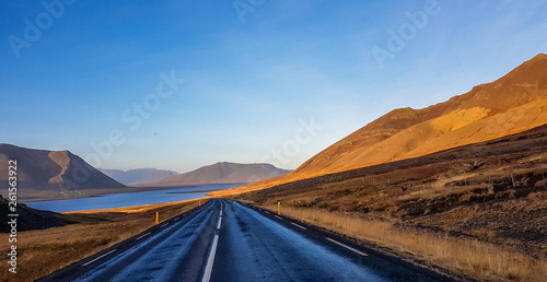An endless road through the lowlands. Both sides of the road are barren. Golden hour  sky is full of colors. Empty road  not a single car passing by. To the side a calm water of a fjord.