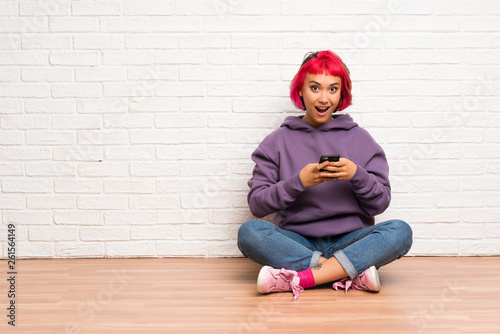 Young woman with pink hair sitting on the floor surprised and sending a message © luismolinero