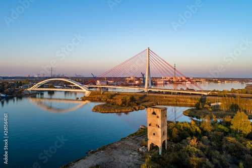 Gdansk, Poland. Modern highway cable-stayed bridge, railway suspension bridge over Dead Vistula river and old Bartos Tower (Wieża Bartosa). Aerial view. Sunset light. Northern Port in the background