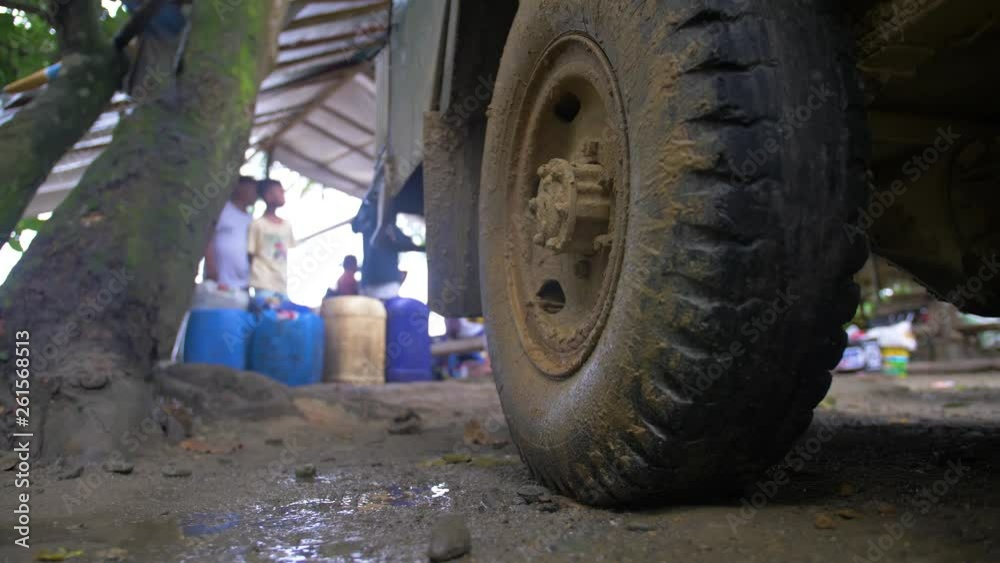 Close up of muddy truck wheel and tire as men gather in the background under a metal roof near blue containers. SLIDE RIGHT. Close up of muddy truck wheel and tire as men gather in the background under a metal roof near blue containers. SLIDE RIGHT.