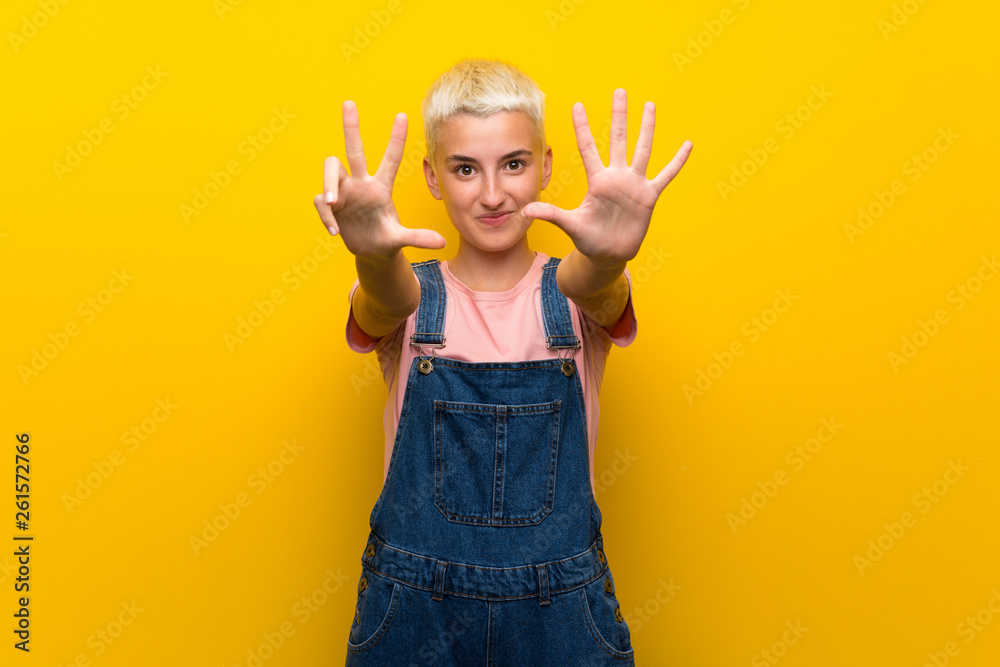 Teenager girl with overalls on yellow background counting eight with fingers