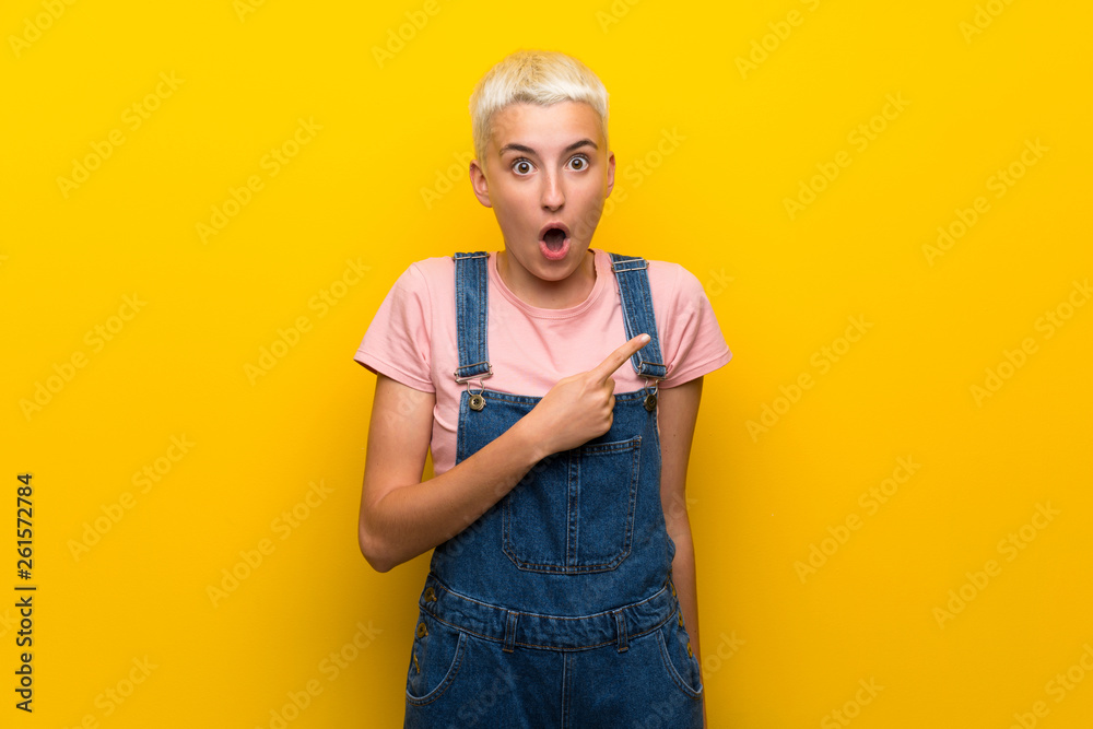 Teenager girl with overalls on yellow background surprised and pointing side Teenager girl with overalls on yellow background surprised and pointing side