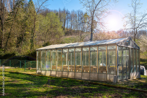 Backlit view of the exterior of a glasshouse in a green lawn with a woody hill in the background and lens flares, Piedmont, Italy