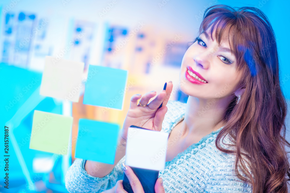 Business woman is engaged in planning. Colored stickers on the glass wall. The girl in the office is planning a meeting. Friendly smile. Brunette makes notes on colored stickers. Business woman is engaged in planning. Colored stickers on the glass wall. The girl in the office is planning a meeting. Friendly smile. Brunette makes notes on colored stickers.