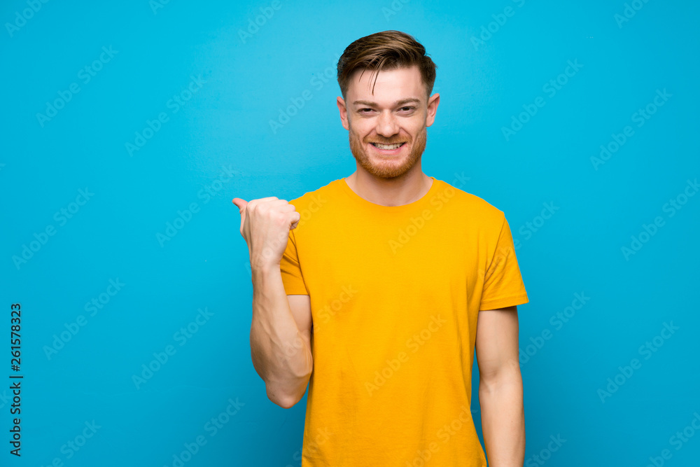 Redhead man over blue wall pointing to the side to present a product Redhead man over blue wall pointing to the side to present a product