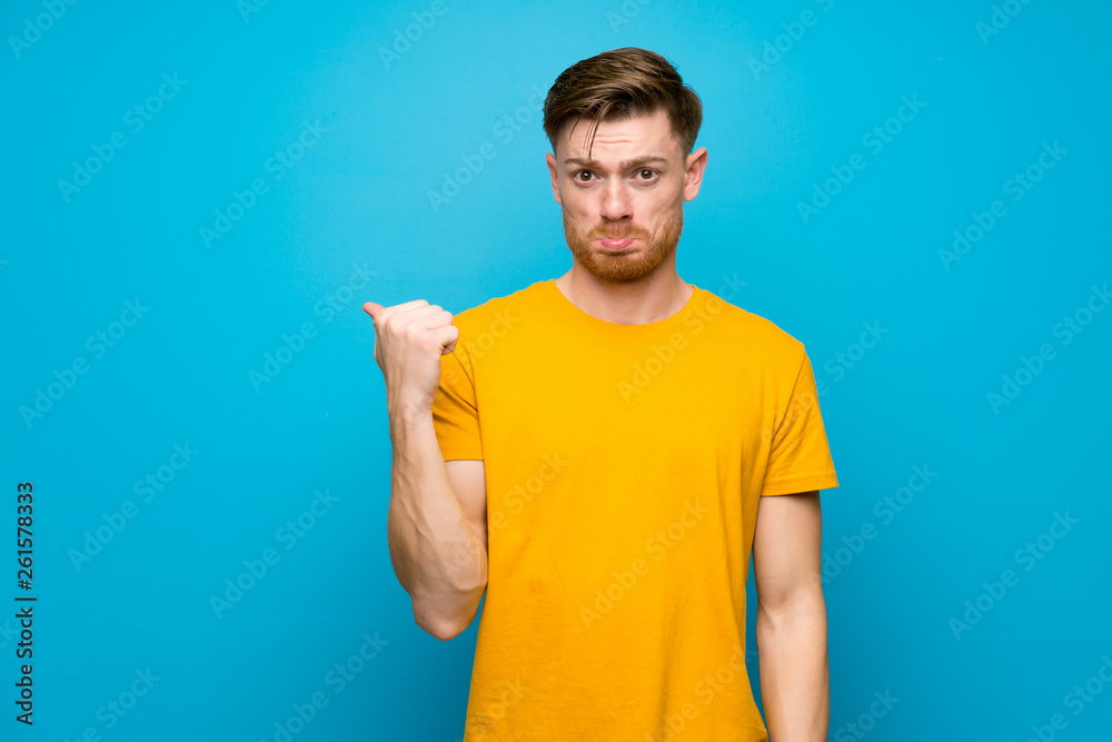 Redhead man over blue wall unhappy and pointing to the side Redhead man over blue wall unhappy and pointing to the side
