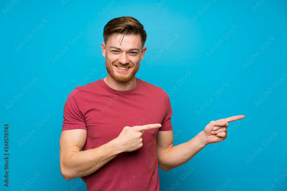 Redhead man on isolated blue wall pointing finger to the side