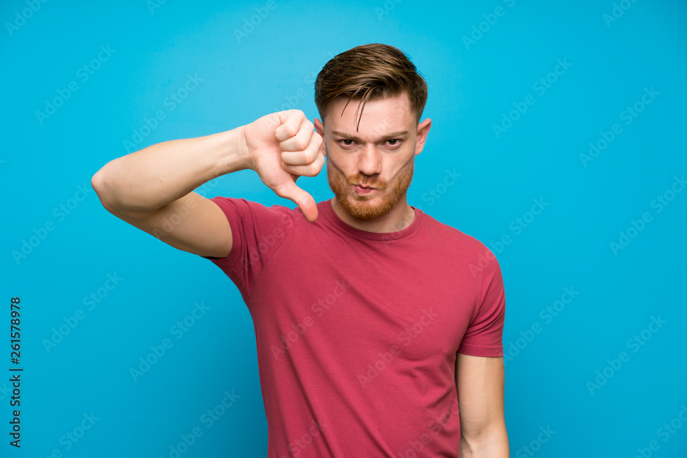 Redhead man on isolated blue wall showing thumb down sign Redhead man on isolated blue wall showing thumb down sign