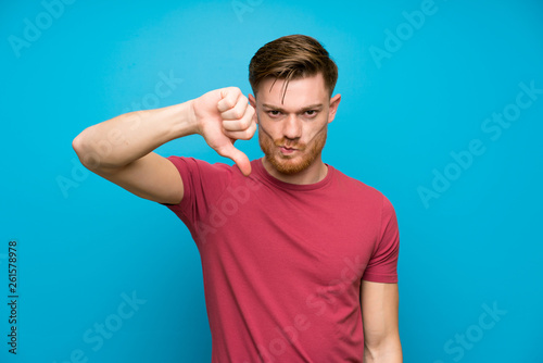 Redhead man on isolated blue wall showing thumb down sign