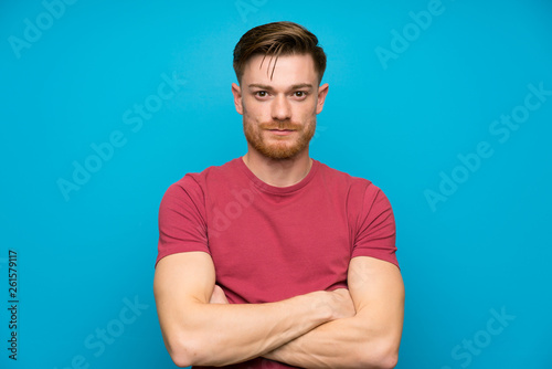 Redhead man on isolated blue wall keeping arms crossed