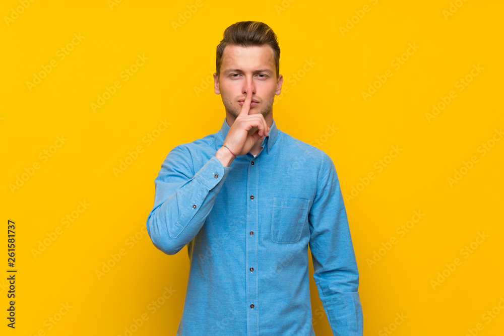 Blonde man over isolated yellow wall showing a sign of silence gesture putting finger in mouth Blonde man over isolated yellow wall showing a sign of silence gesture putting finger in mouth