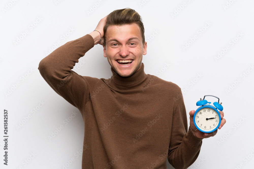 Blonde man over isolated white wall restless because it has become late and holding vintage alarm clock Blonde man over isolated white wall restless because it has become late and holding vintage alarm clock