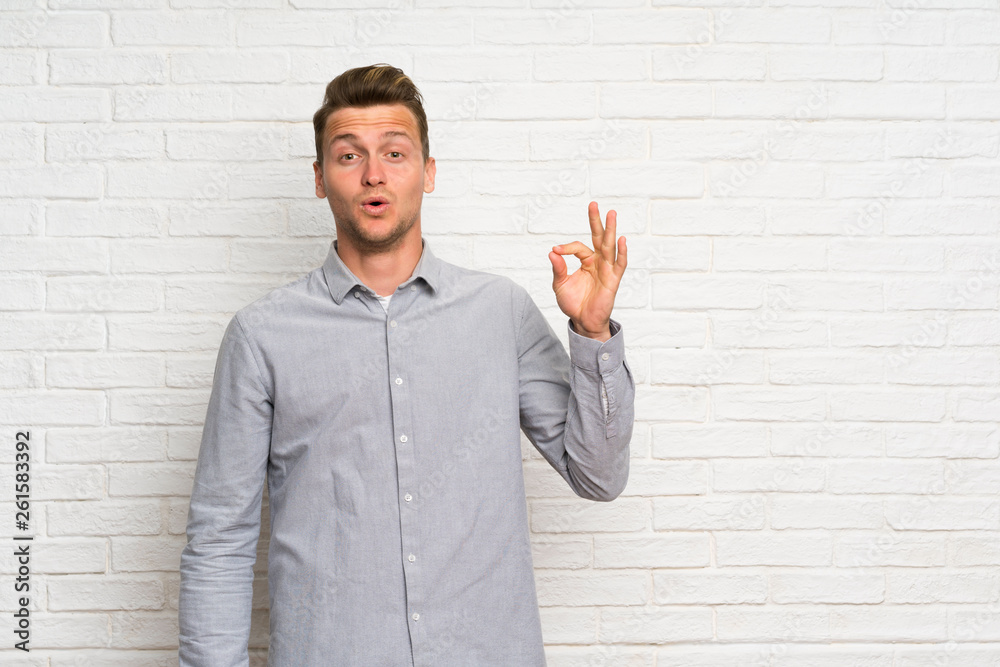 Blonde man over white brick wall surprised and showing ok sign Blonde man over white brick wall surprised and showing ok sign