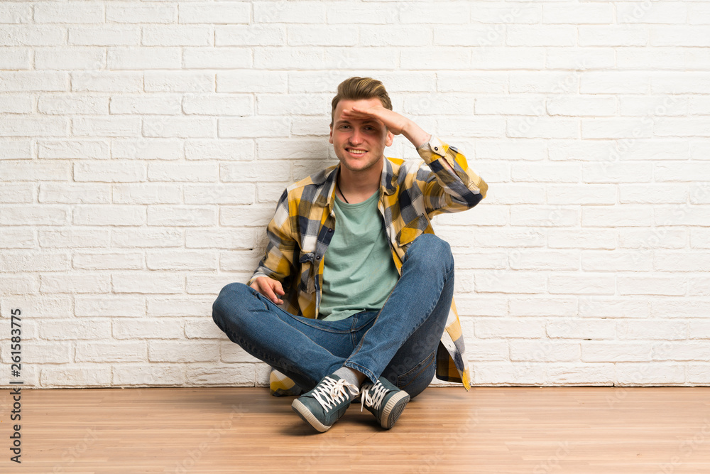Blonde man sitting on the floor looking far away with hand to look something Blonde man sitting on the floor looking far away with hand to look something