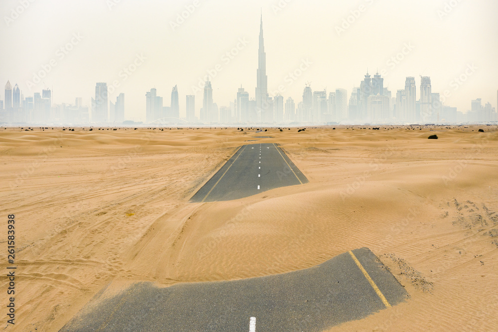 Aerial view of a deserted road covered by sand dunes in the middle of the Dubai desert. Dubai skyline with the Burj Khalifa surrounded by fog in the background. Dubai, United Arab Emirates.