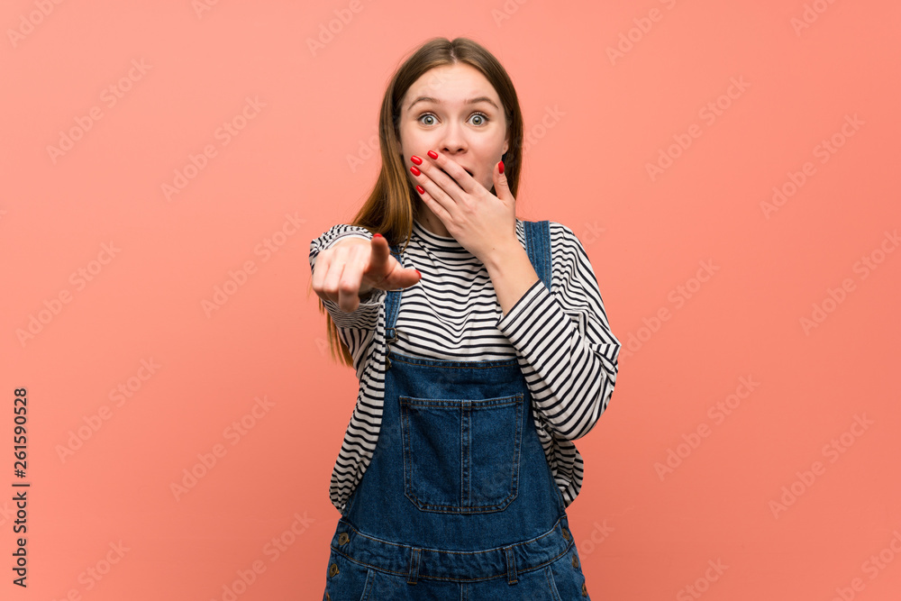 Young woman with overalls over pink wall pointing with finger at someone and laughing Young woman with overalls over pink wall pointing with finger at someone and laughing
