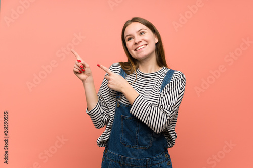 Young woman with overalls over pink wall pointing with the index finger and looking up