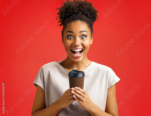 Joyful girl holds cup of hot coffee. Photo of african american girl wears casual outfit on red background. Emotions and pleasant feelings concept.