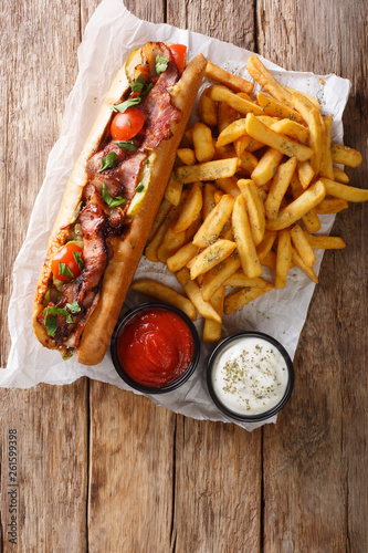 Rustic style hot dog with bacon and vegetables served with french fries and sauces close-up. Vertical top view