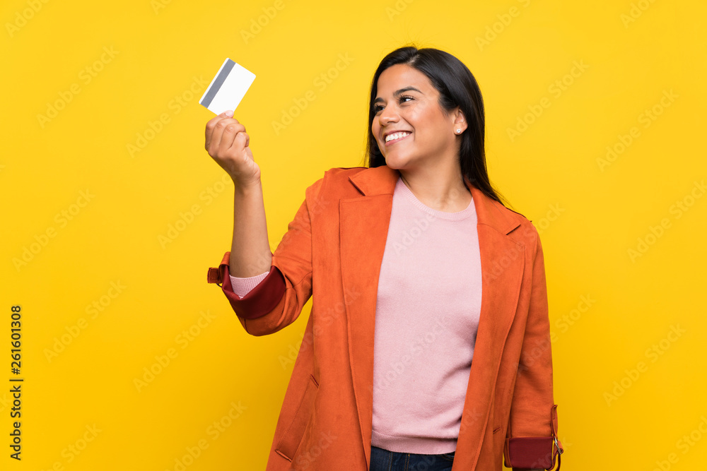 Young Colombian girl over yellow wall holding a credit card and thinking Young Colombian girl over yellow wall holding a credit card and thinking