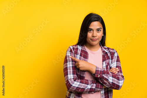 Young Colombian girl over yellow wall pointing finger to the side © luismolinero