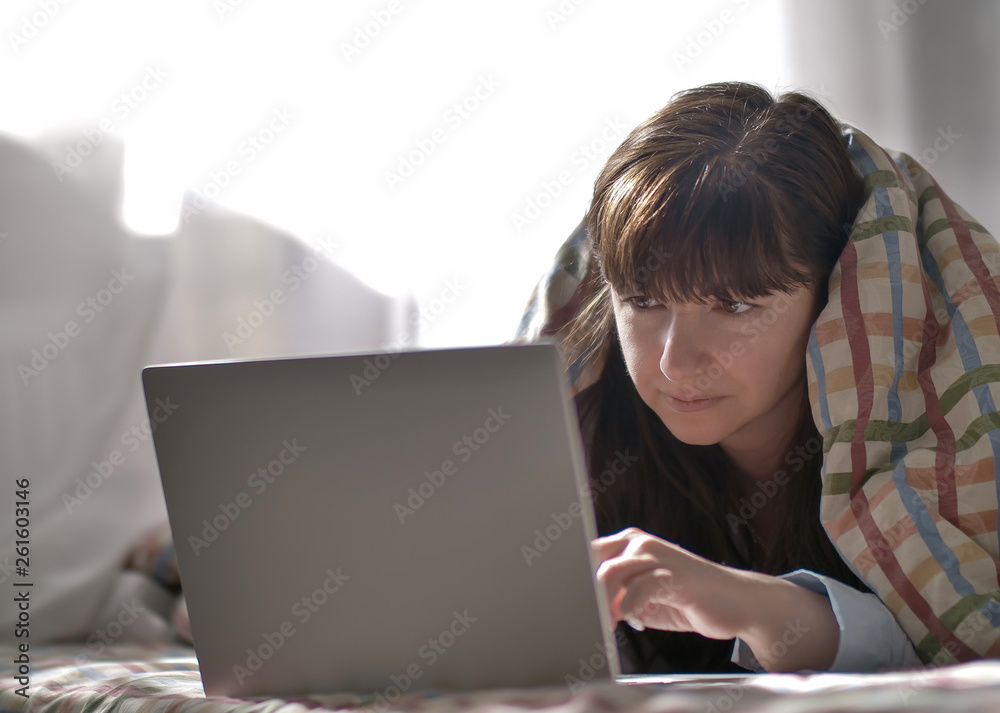 Young brunette woman lies under the covers and looks into the laptop Young brunette woman lies under the covers and looks into the laptop