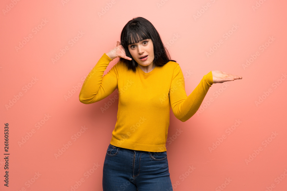 Woman with yellow sweater over pink wall making phone gesture and doubting Woman with yellow sweater over pink wall making phone gesture and doubting