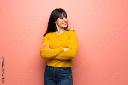 Woman with yellow sweater over pink wall with arms crossed and happy