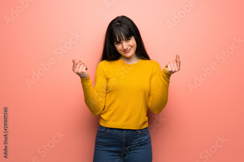 Woman with yellow sweater over pink wall making money gesture