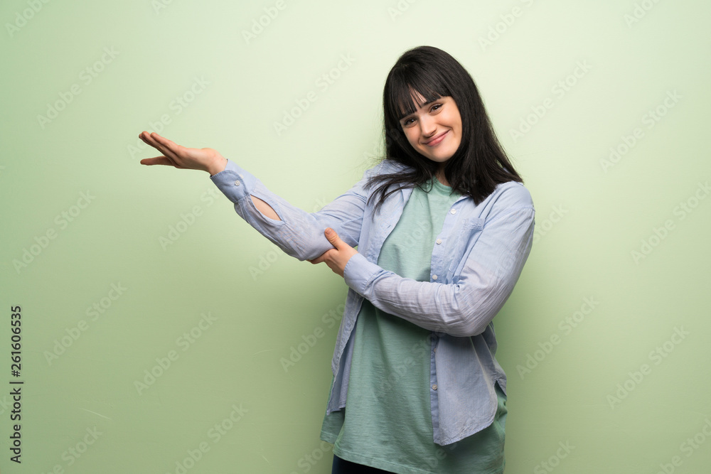Young woman over green wall extending hands to the side for inviting to come Young woman over green wall extending hands to the side for inviting to come