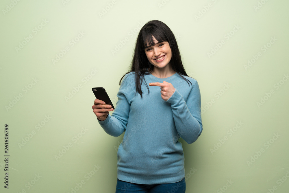 Young woman over green wall happy and pointing the mobile Young woman over green wall happy and pointing the mobile