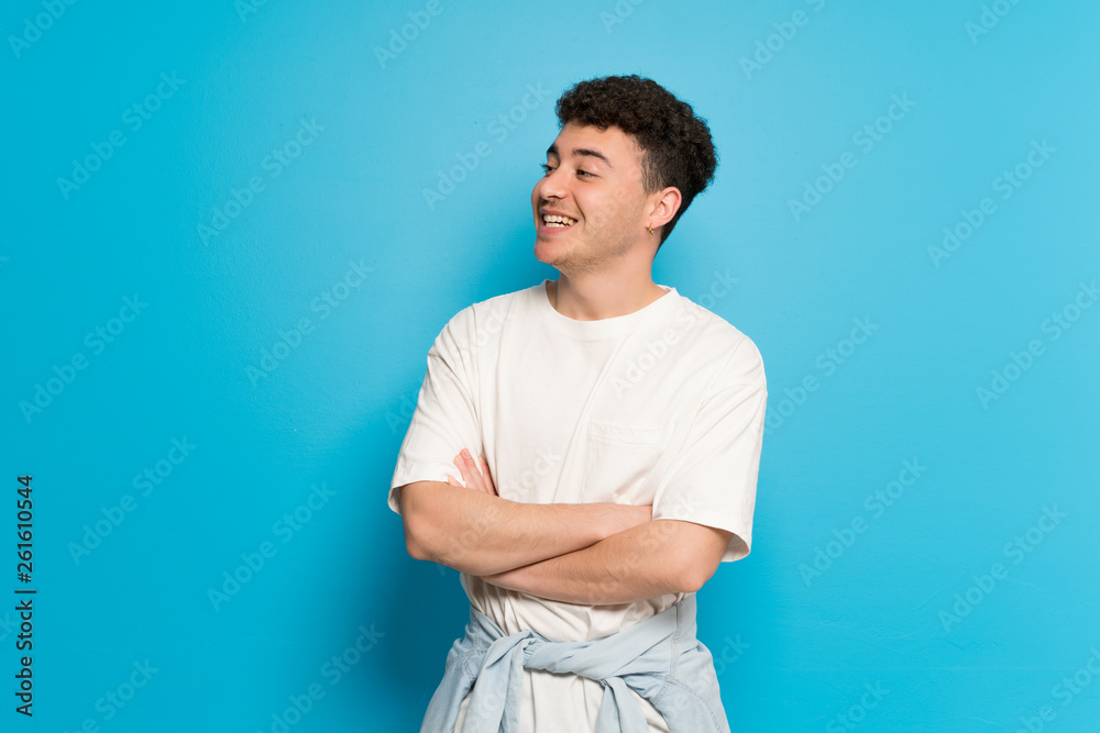 Young man over blue background keeping the arms crossed while smiling Young man over blue background keeping the arms crossed while smiling
