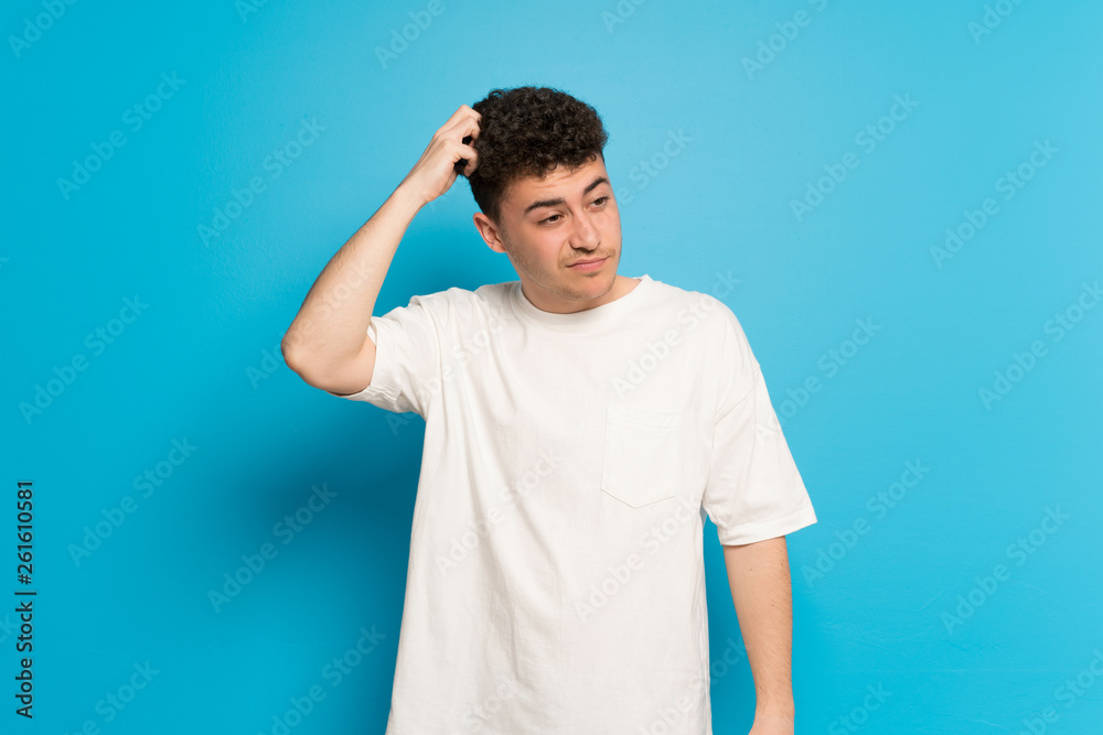 Young man over blue background having doubts while scratching head Young man over blue background having doubts while scratching head