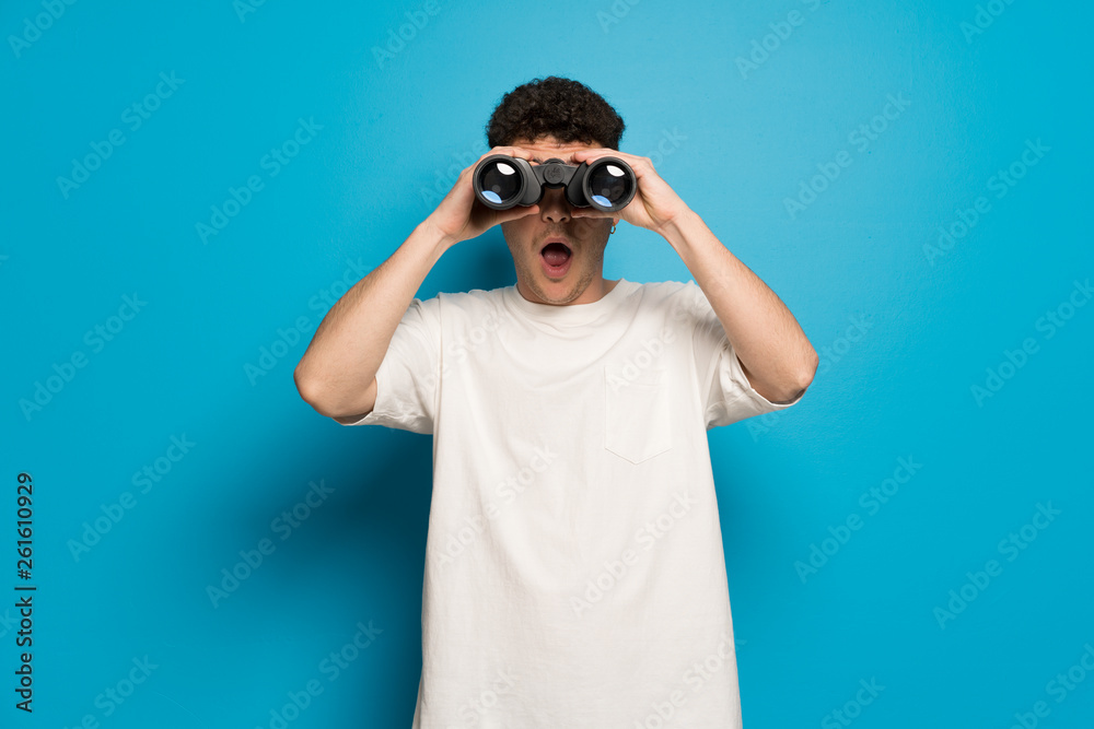Young man over blue background and looking in the distance with binoculars Young man over blue background and looking in the distance with binoculars