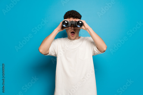 Young man over blue background and looking in the distance with binoculars