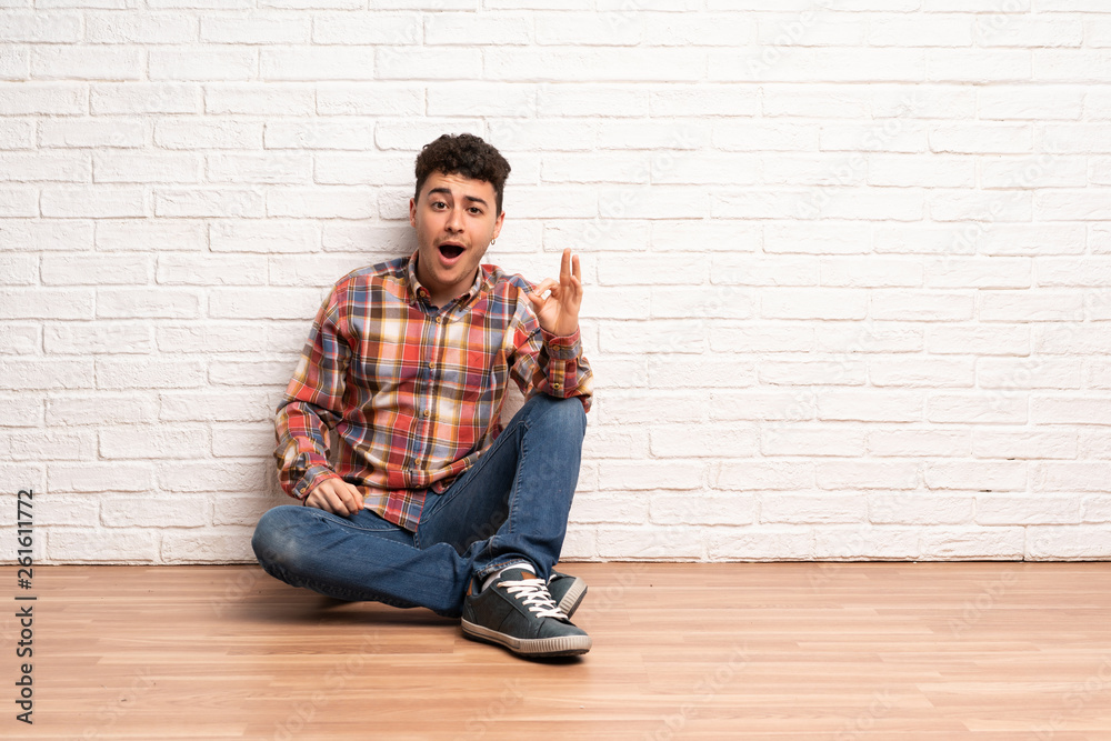Young man sitting on the floor surprised and showing ok sign Young man sitting on the floor surprised and showing ok sign